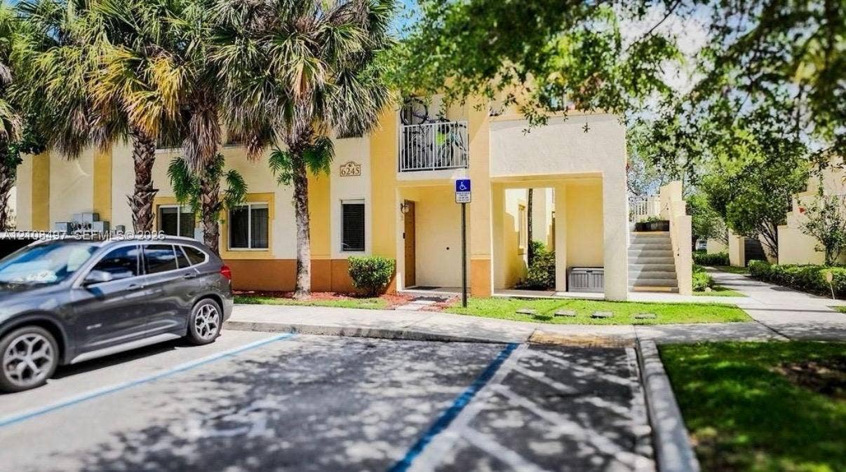 6245 SW 47th Manor, #101, Davie, FL 33314 Exterior facade featuring a two-story stucco finish, a covered balcony with metal railings, a concrete stairway, and mature palm trees