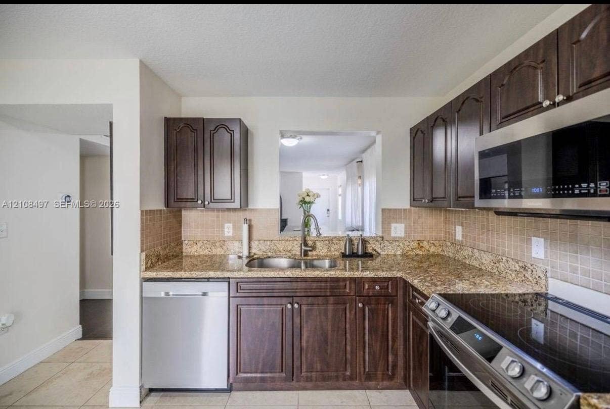 6245 SW 47th Manor, #101, Davie, FL 33314 Kitchen featuring dark-finish wood cabinetry, stone-surface countertops, square tile backsplash, and stainless steel appliances including a built-in microwave and dishwasher