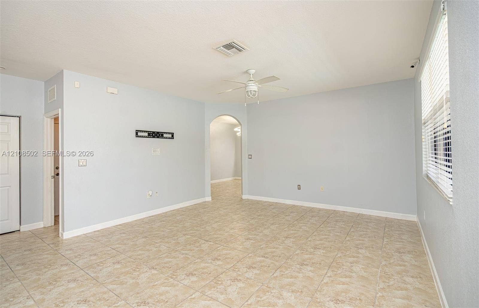 180 NE 26th Avenue, Homestead, FL 33033 Neutral-toned interior space featuring light-colored tile flooring, an arched doorway, a ceiling fan with integrated lighting, and horizontal window blinds