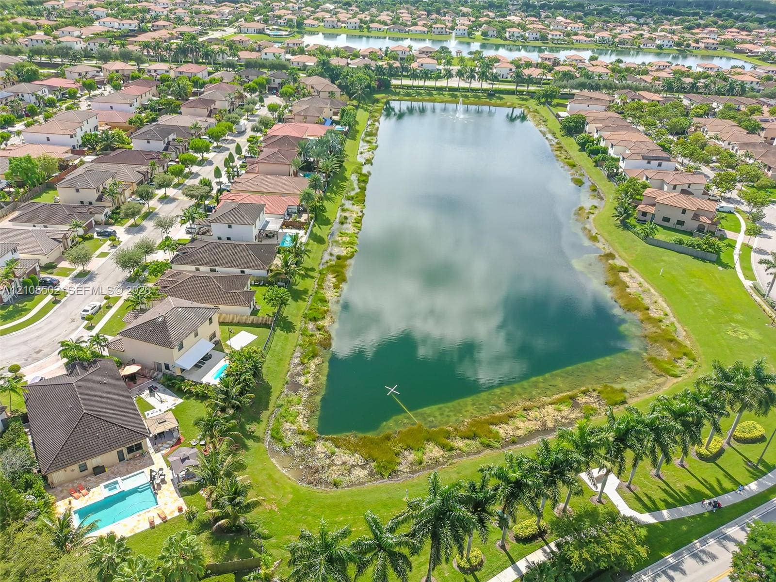 Aerial perspective featuring a multi-story residence with a paved pool deck, rectangular swimming pool, manicured lawn, and proximity to a large central lake