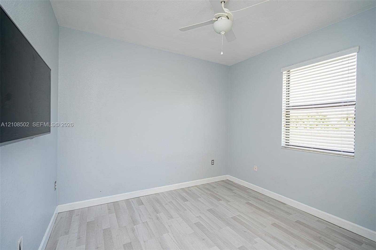 Neutral wall-toned room featuring wood-finish flooring, a white ceiling fan, and a window with horizontal blinds