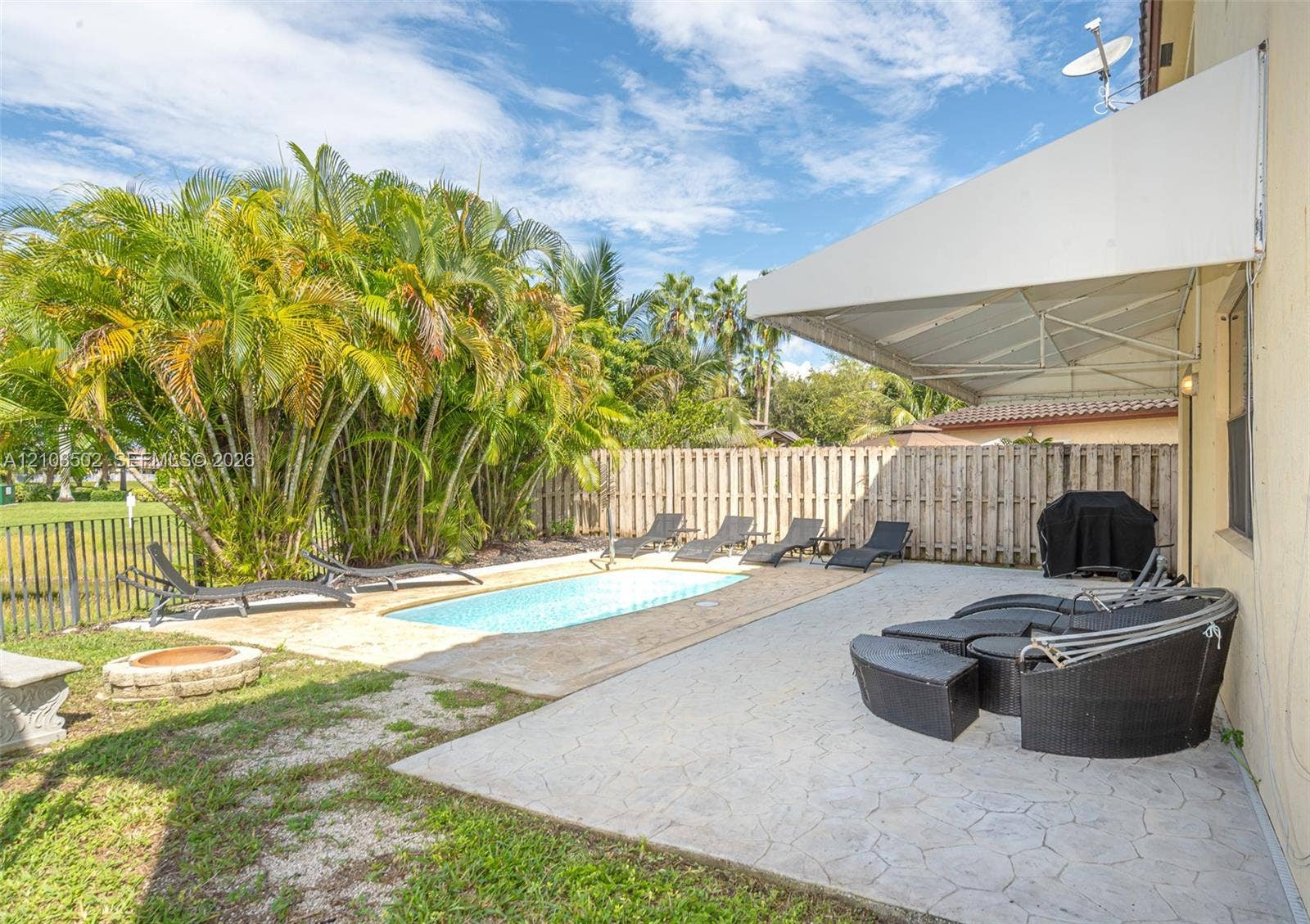 180 NE 26th Avenue, Homestead, FL 33033 Backyard featuring a concrete-paver patio, retractable fabric awning, in-ground swimming pool, stone-finish fire pit, and wood-style privacy fencing