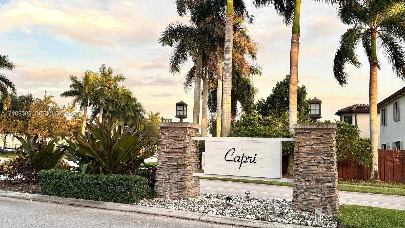 180 NE 26th Avenue, Homestead, FL 33033 Community entrance featuring stacked stone pillars, decorative post-top lighting, and mature palm tree landscaping