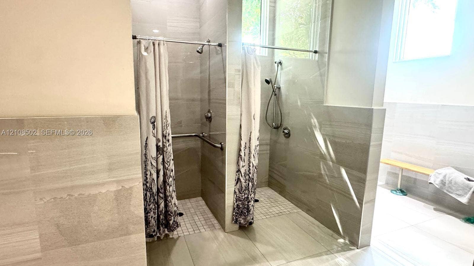 Dual walk-in shower stalls featuring large-format neutral wall tiling, mosaic floor tile, chrome shower fixtures with handheld sprayers, and a built-in wall bench