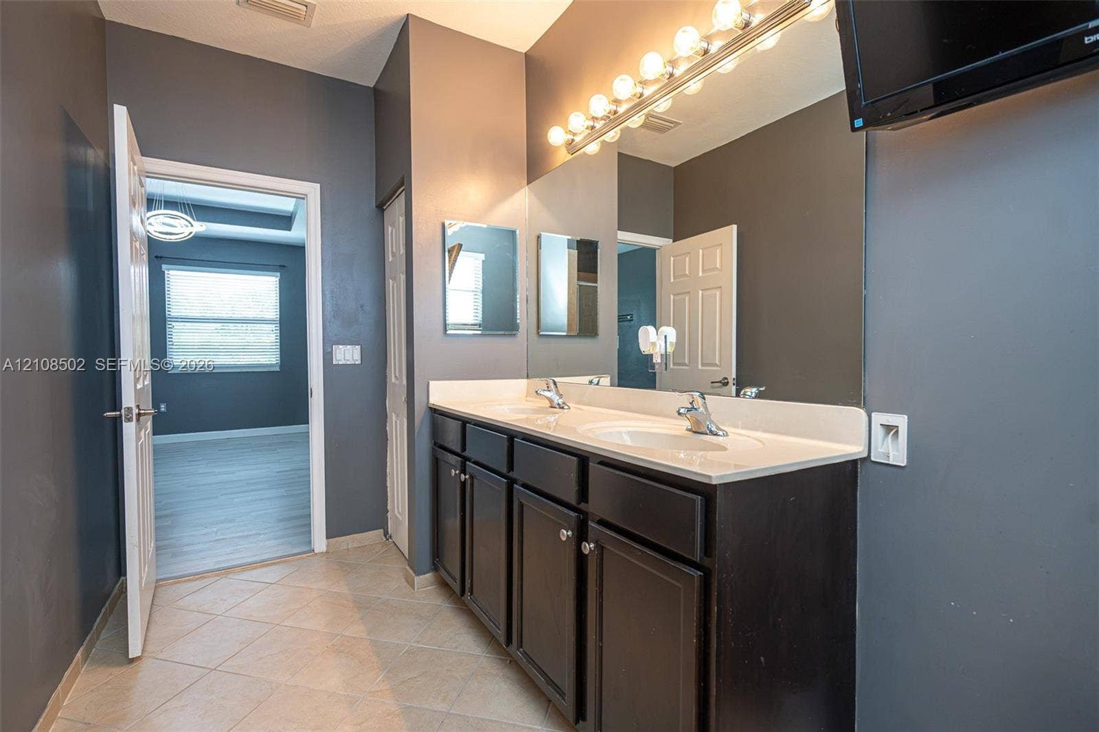 180 NE 26th Avenue, Homestead, FL 33033 Dual-sink vanity with a solid-surface countertop, dark cabinetry, full-width mirror, and multi-bulb Hollywood-style lighting, complemented by tile flooring