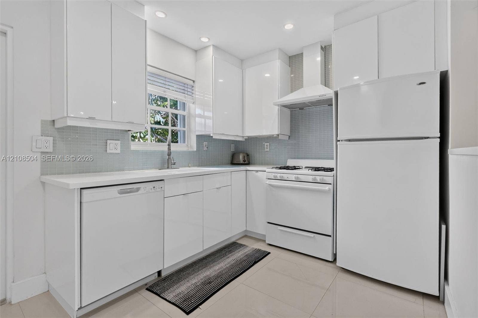 Kitchen featuring flat-panel white cabinetry, light-toned tile flooring, light blue glass tile backsplash, white countertops, and a suite of white appliances