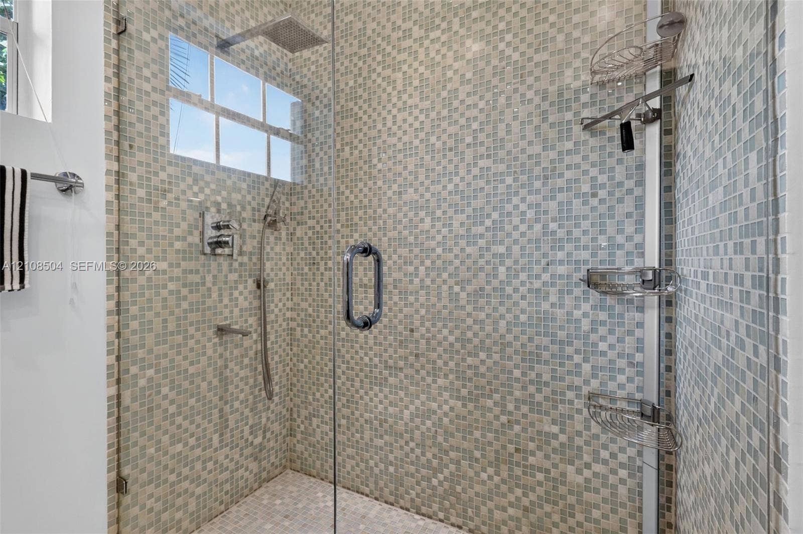 Walk-in shower featuring a frameless glass door, multi-tone mosaic wall tile, overhead rain shower head, handheld shower attachment, and integrated corner shelving