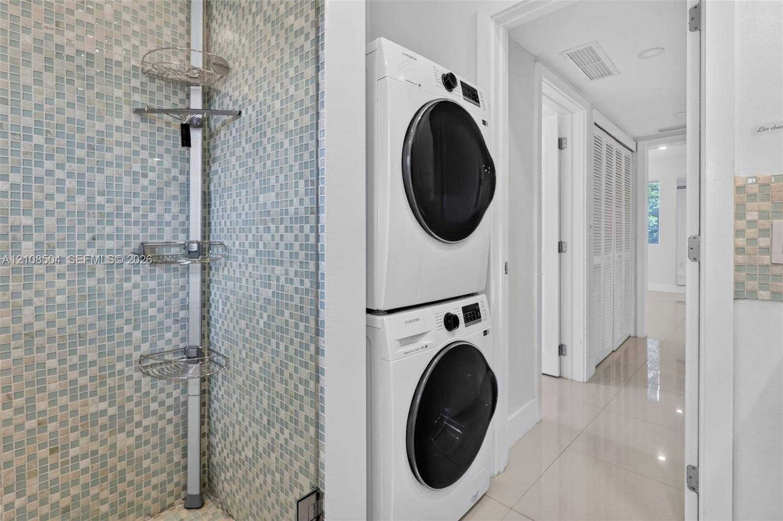 Laundry area featuring stacked front-load washer and dryer, tiled wall with integrated shelving, and polished neutral-tone flooring