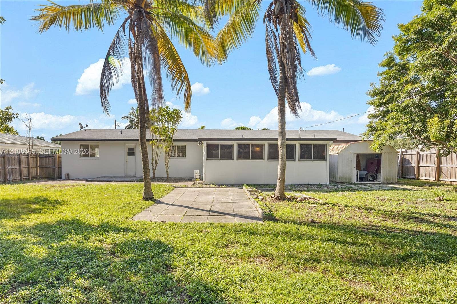 Spacious backyard featuring a grass lawn, paved patio, mature palm trees, detached storage structure, and wood privacy fencing