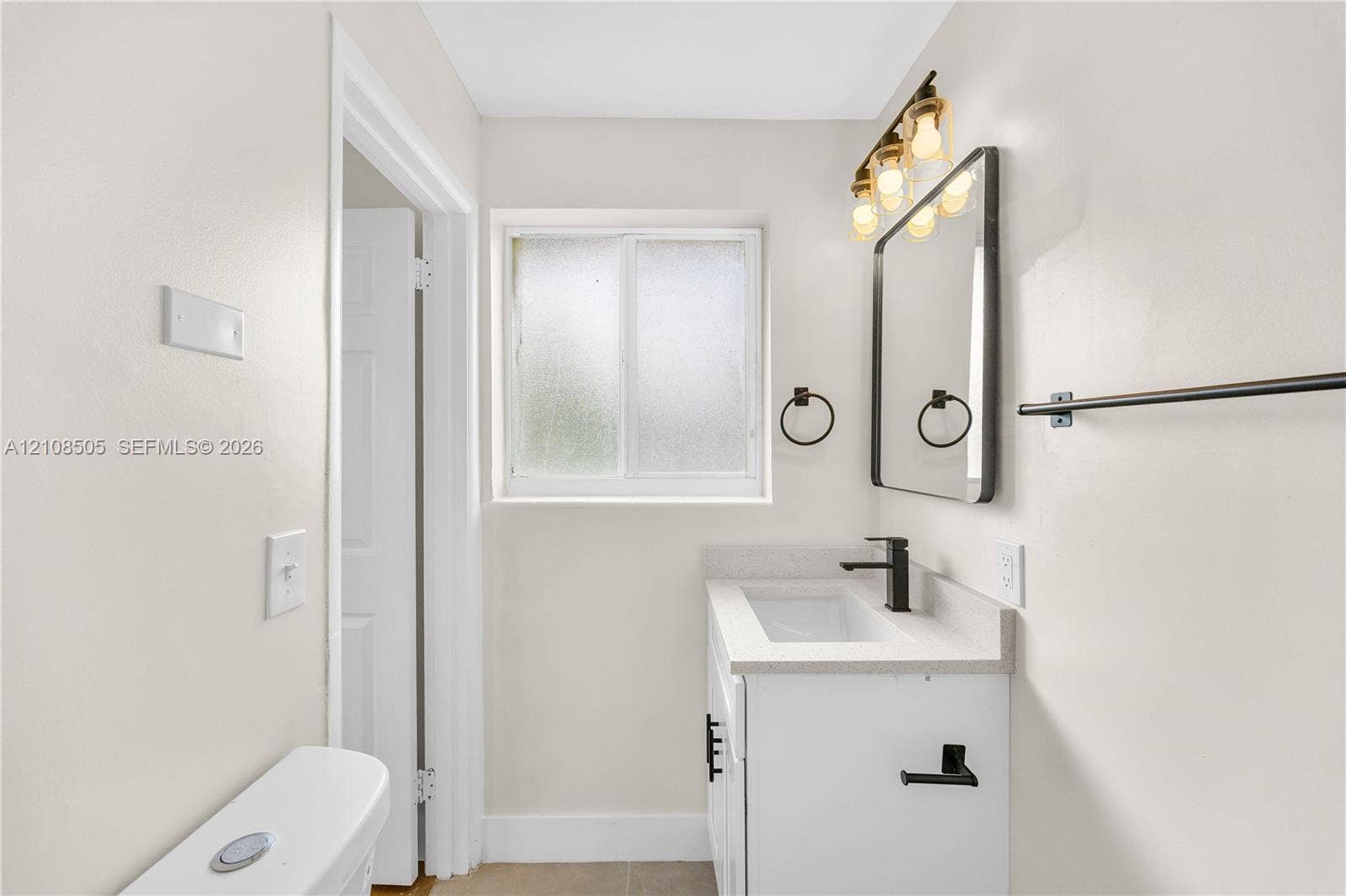 White vanity with a neutral solid-surface countertop, rectangular integrated basin, matte black faucet, and matching black hardware, complemented by a rectangular mirror with a black frame and multi-bulb vanity light fixture
