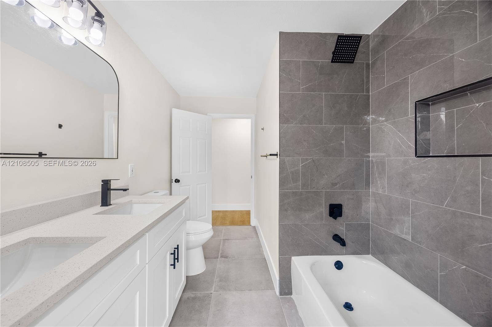 Double vanity with a stone-finish countertop, matte black plumbing fixtures, arched wall mirror, and a bathtub with full-height stone-finish tile surround and built-in recessed niche