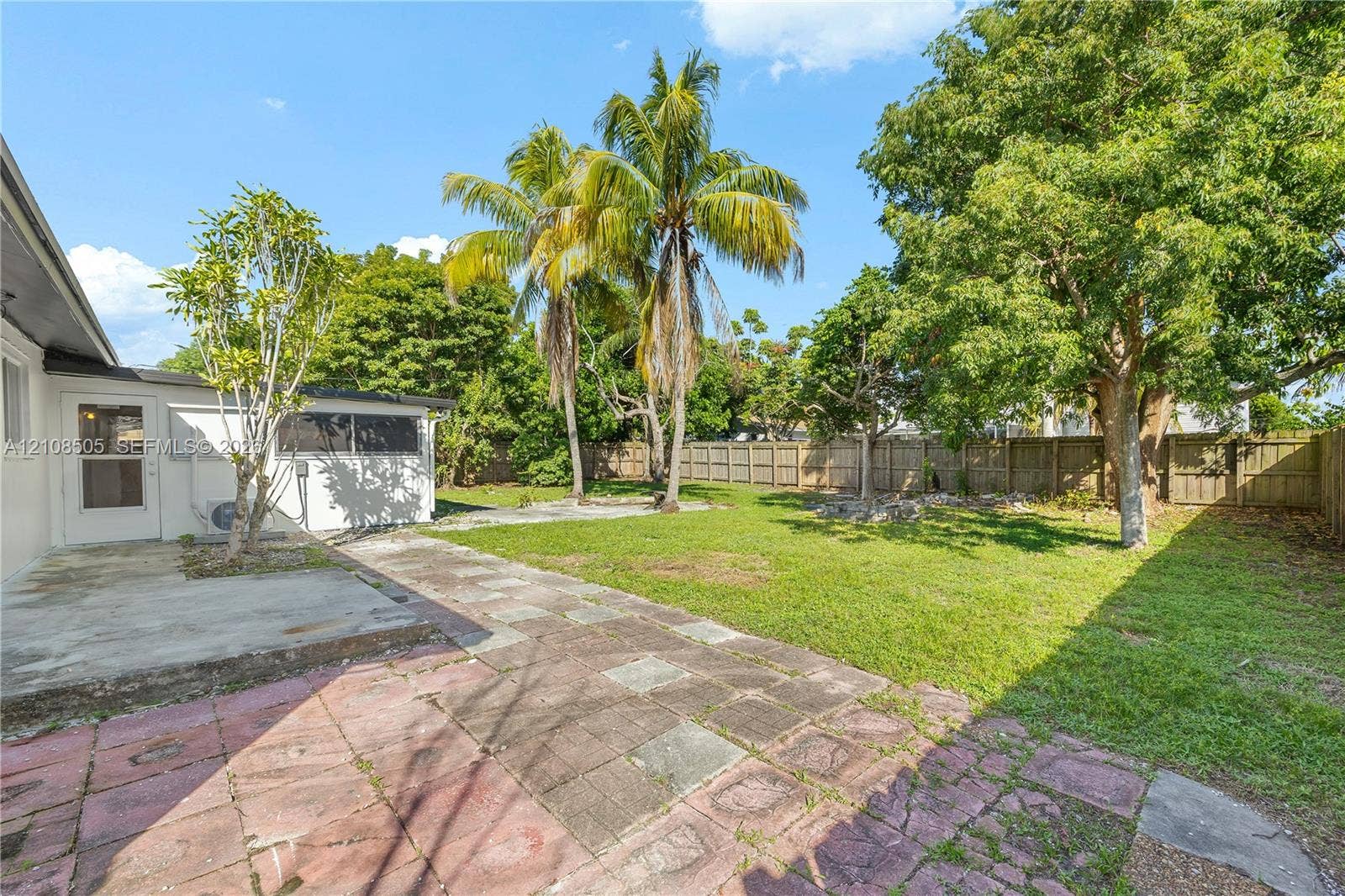 Backyard featuring a paved stone patio, expansive lawn, mature palm and shade trees, and a perimeter wood privacy fence