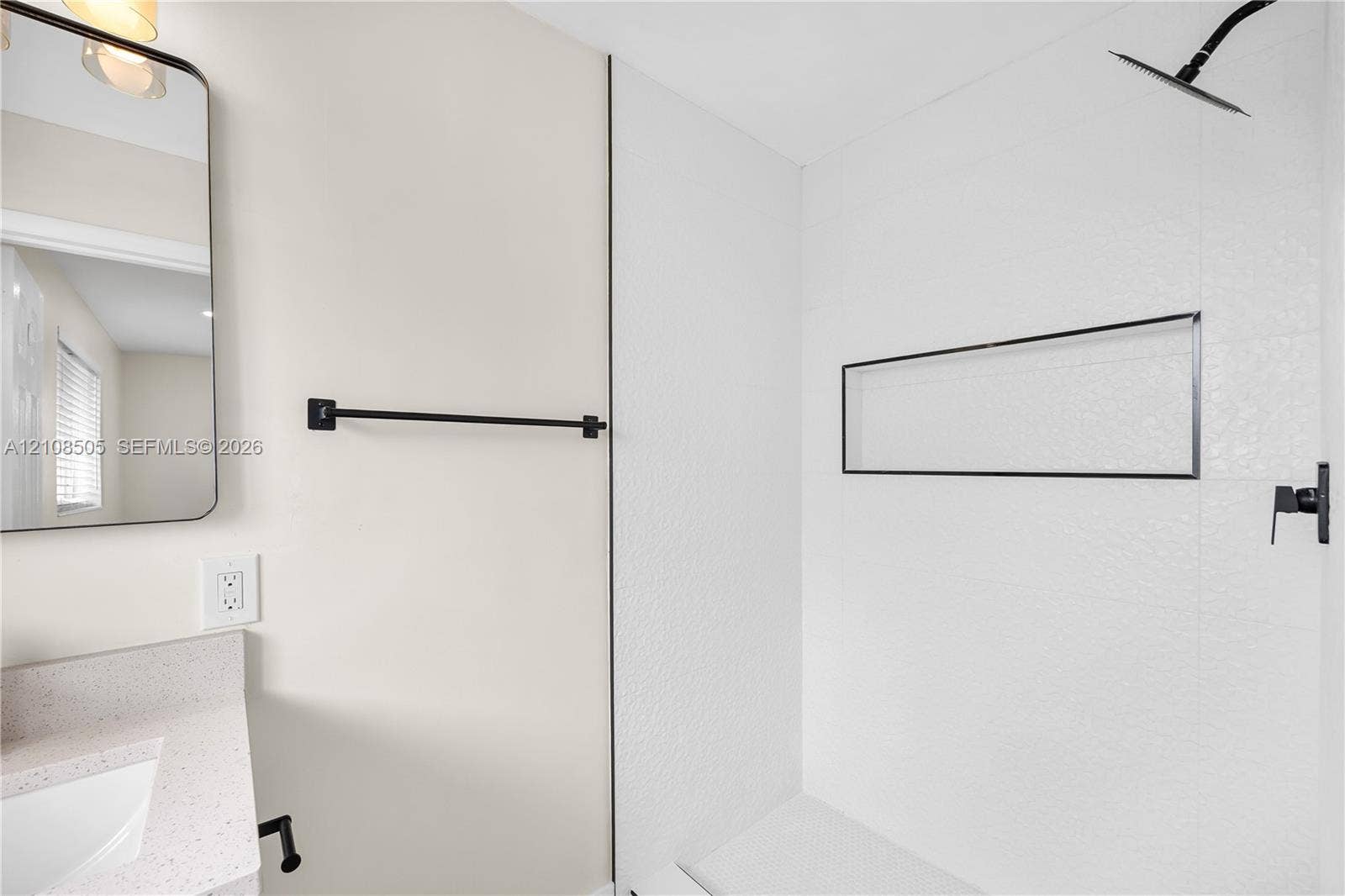 Bathroom featuring a walk-in shower with textured tile surround and built-in recessed shelf, matte black wall-mounted shower head and fixtures, stone-finish countertop with integrated sink, and an arched wall-mounted mirror