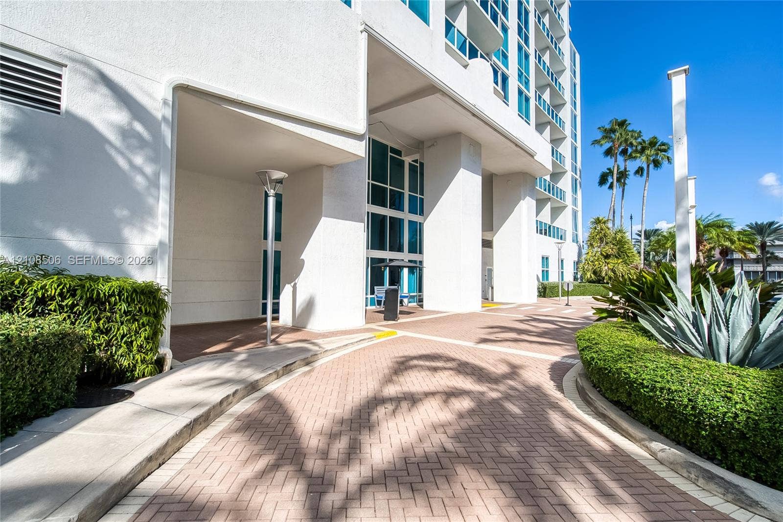 High-rise exterior featuring concrete structural pillars, floor-to-ceiling glass windows, a brick paver driveway, and manicured landscaping