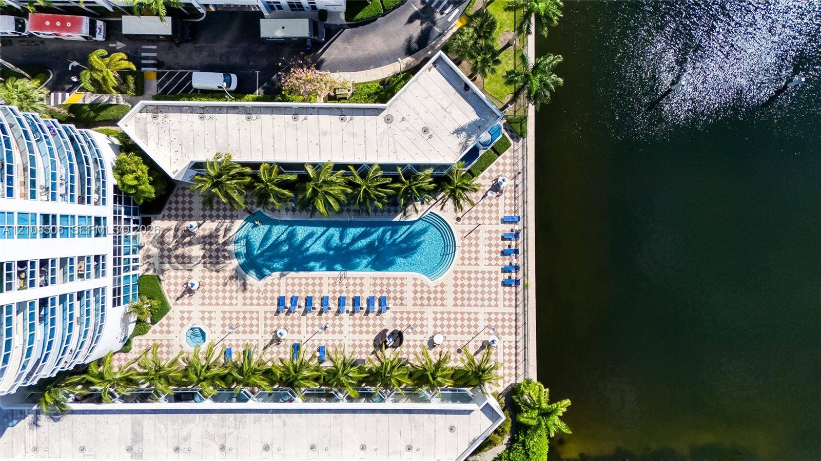 Aerial perspective of a waterfront pool deck featuring a geometric paved patio, swimming pool, in-ground spa, and mature palm trees