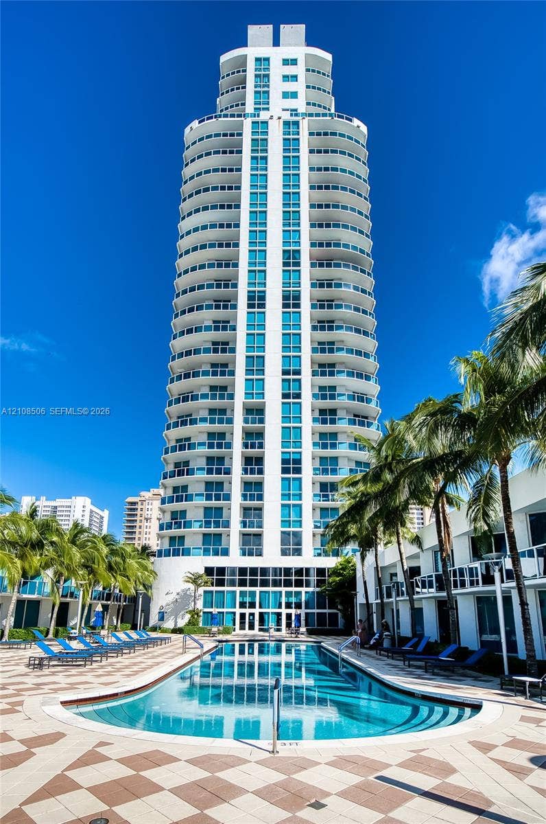High-rise residential tower featuring wrap-around balconies and floor-to-ceiling glass windows, accented by a centered ground-level pool with tile decking and surrounding palm trees