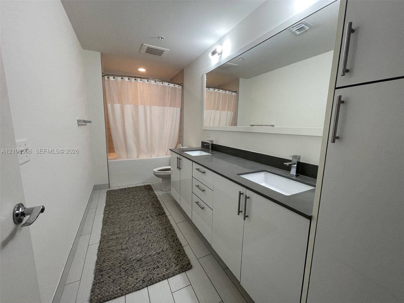 Bathroom featuring a double-sink vanity with white cabinetry, dark solid-surface countertops, a full-width mirror, and tile flooring