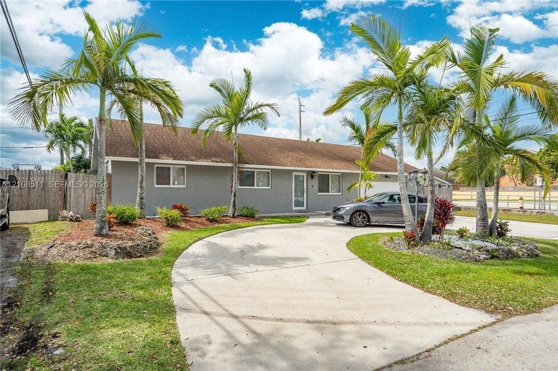 Miramar, Miramar, FL 33025 Single-story exterior featuring a concrete driveway, established palm trees, landscaped garden beds, and a wood fence