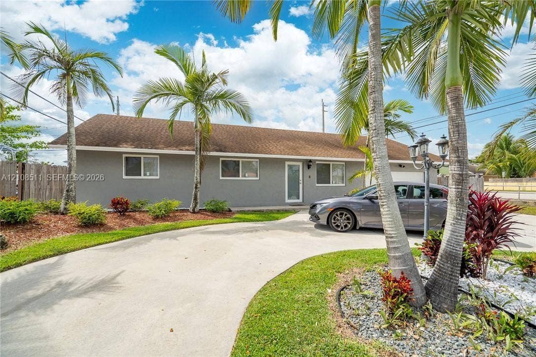 Miramar, Miramar, FL 33025 Single-story exterior featuring a gray stucco facade, arched concrete driveway, multi-pane windows, and landscaped grounds with mature palm trees