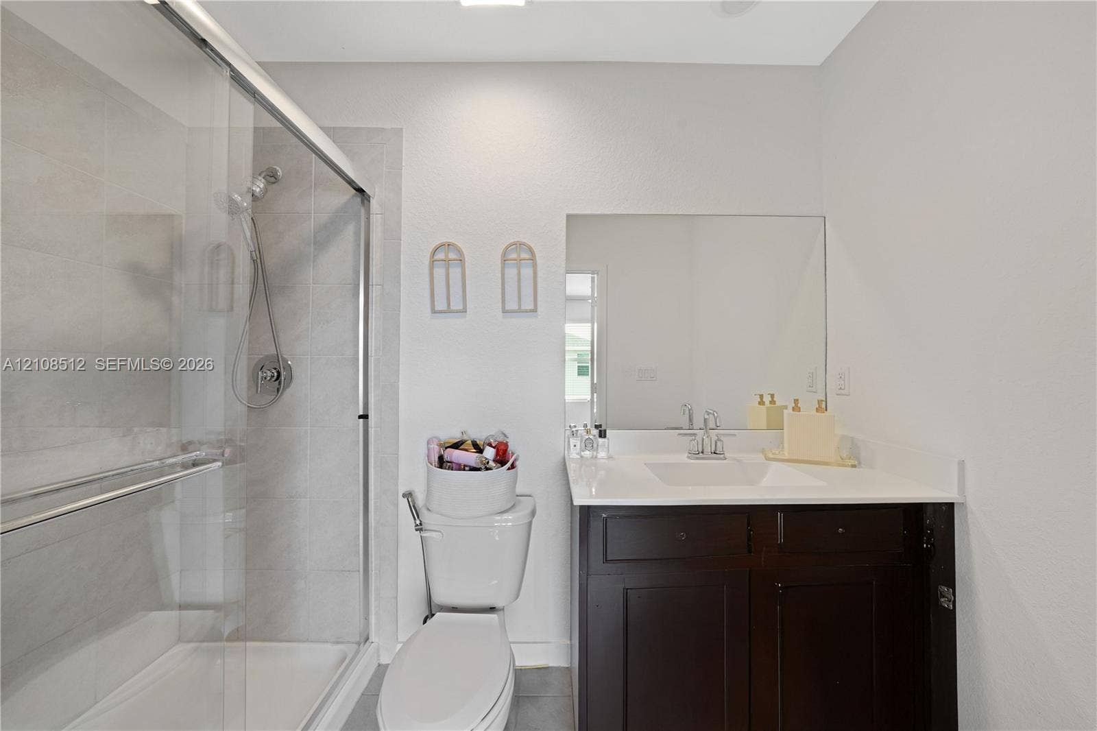 Walk-in shower with sliding glass enclosure and tiled walls, dark-toned vanity cabinet, white solid-surface countertop with integrated basin, and expansive frameless wall mirror