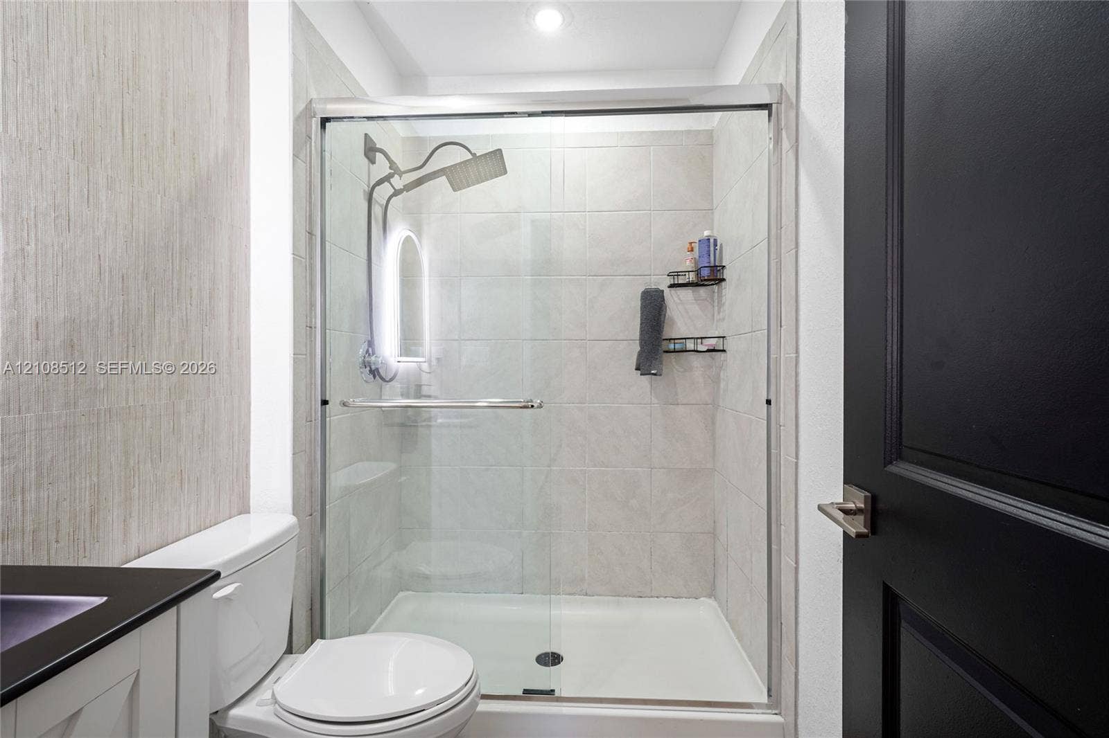 Walk-in shower featuring glass sliding doors, neutral ceramic tile surround, and a rain-style showerhead, complemented by a matte black interior door with matching hardware