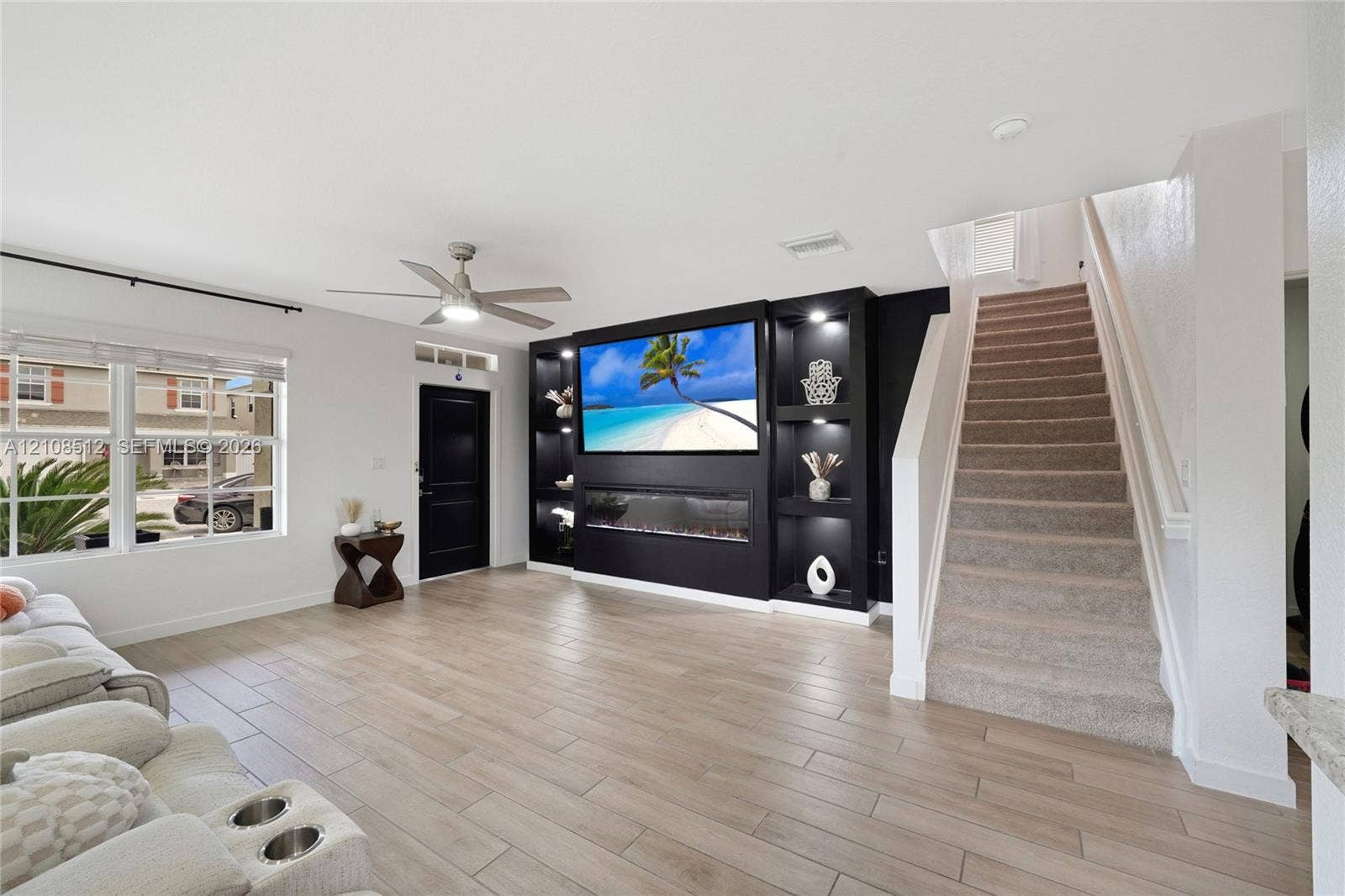 Living area featuring wood-finish flooring, a custom black built-in wall unit with integrated shelving and an electric fireplace, a recessed ceiling fan, and a staircase with carpeted treads