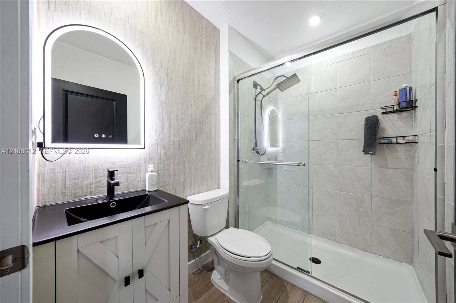 Bathroom featuring a vanity with a dark countertop and integrated sink, arch-top lighted mirror, wood-finish flooring, and a walk-in shower with a glass enclosure and tiled walls