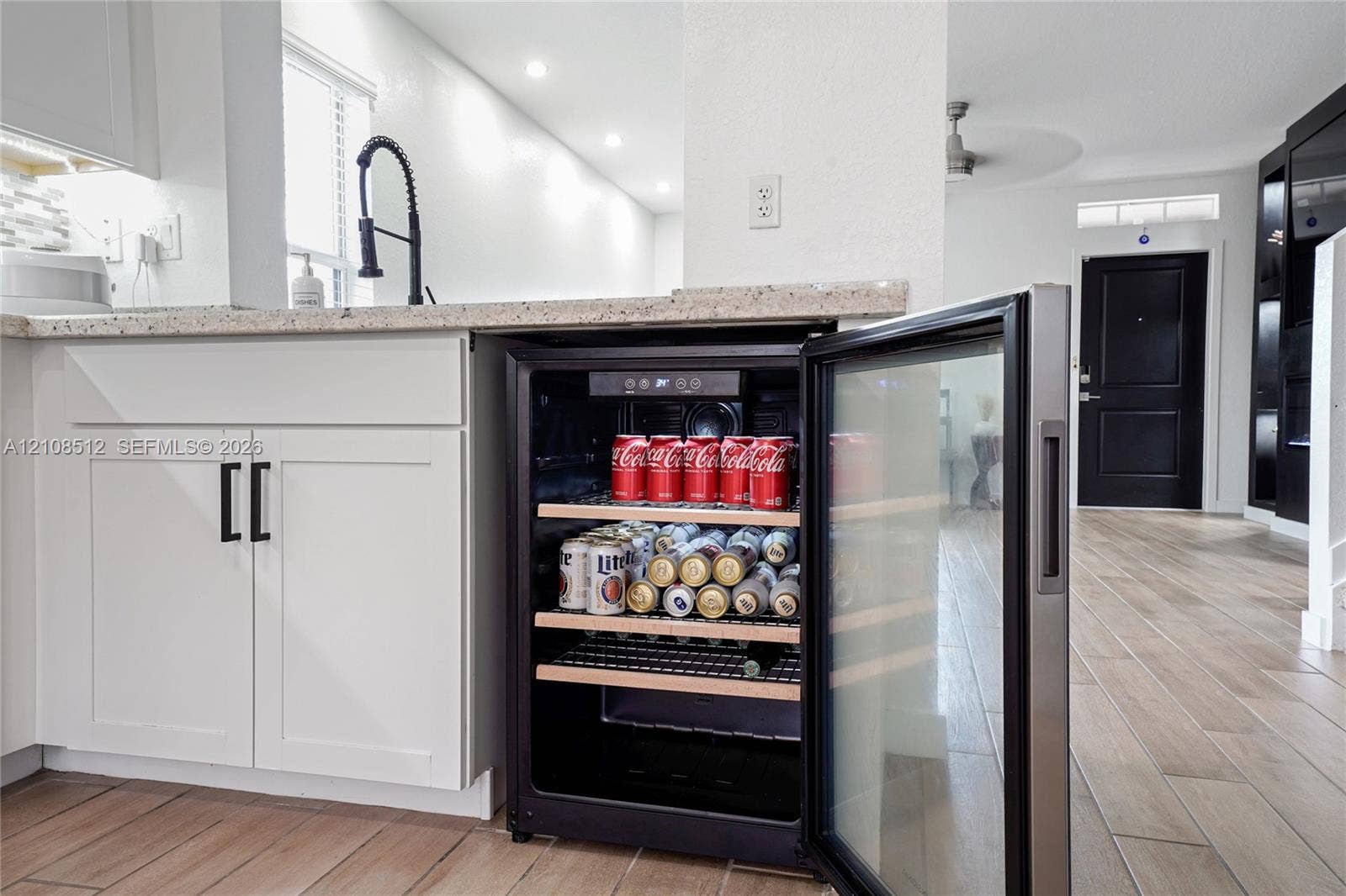 Built-in under-counter beverage refrigerator featuring a glass door and wooden shelving, complemented by white cabinetry, light-toned stone-finish countertops, and wood-finish flooring