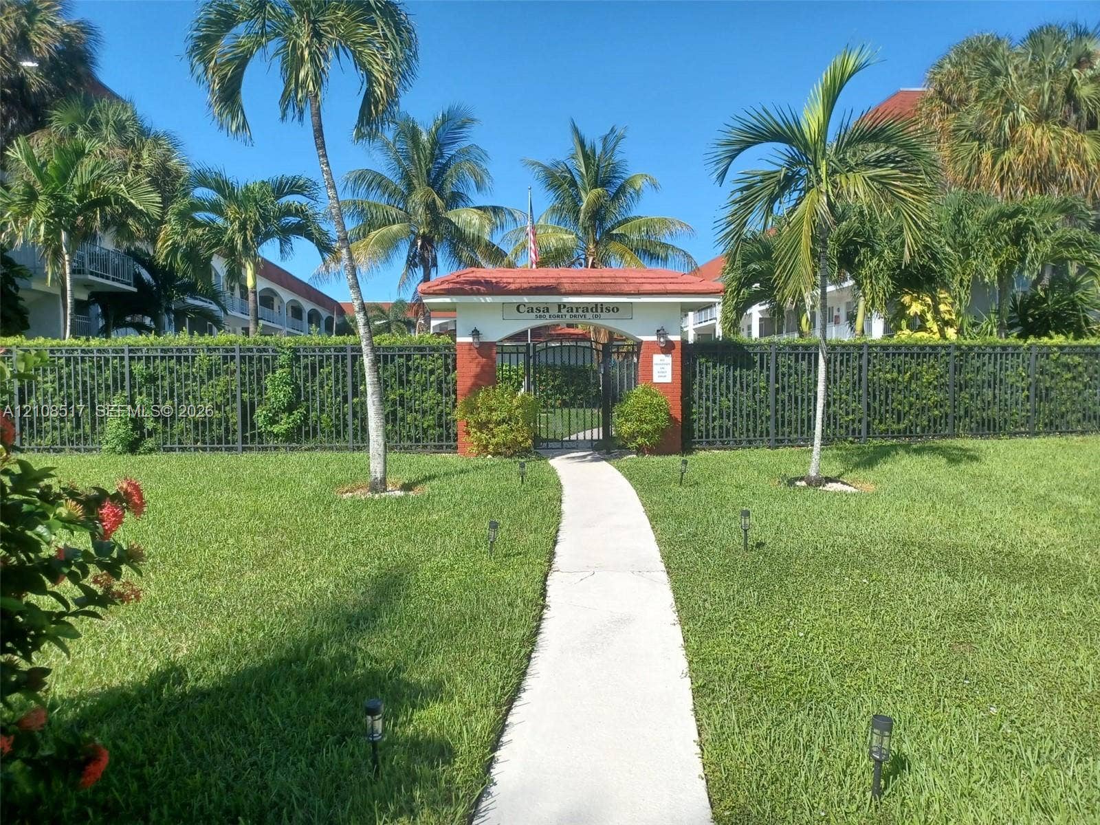541 Blue Heron Drive, #204, Hallandale Beach, FL 33009 Gated entry featuring a terracotta-tiled archway, manicured lawn, perimeter fencing, and mature palm landscaping