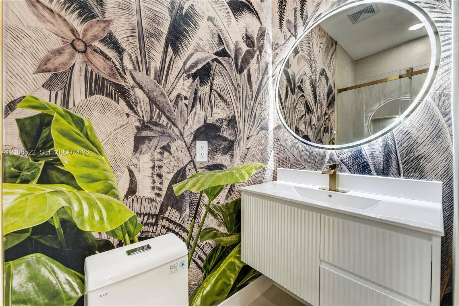 Floating white vanity with vertical paneling, integrated sink, brass-finish faucet, circular backlit mirror, and floor-to-ceiling botanical wall treatment