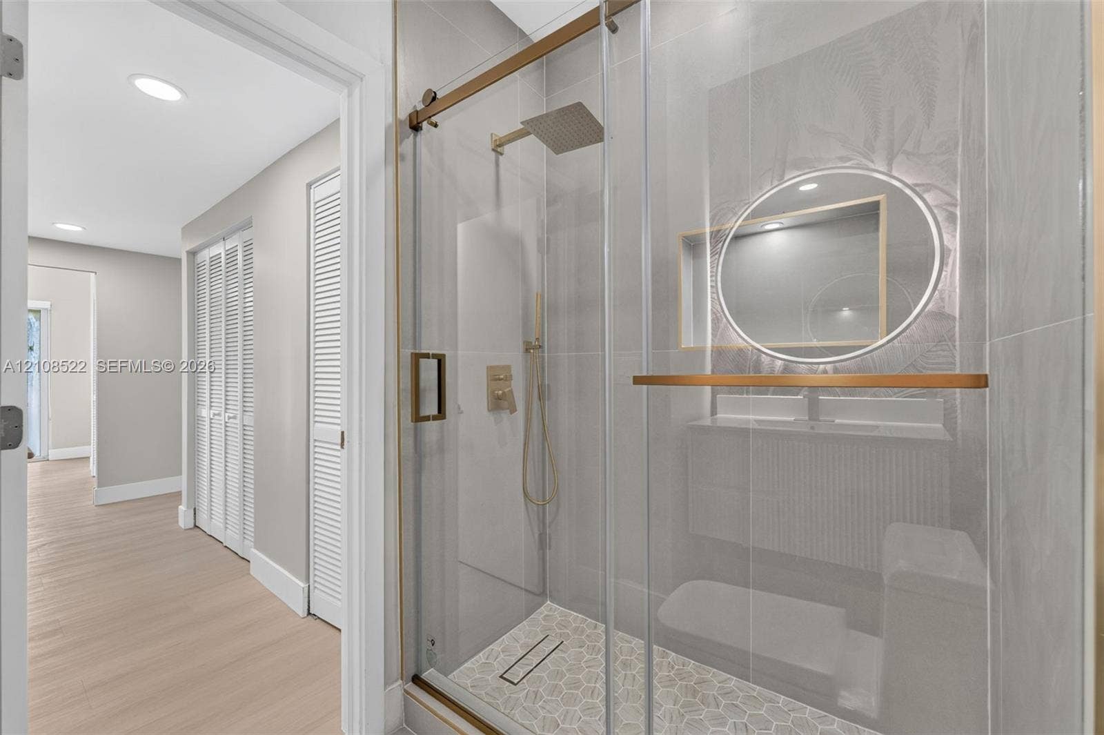 Walk-in shower featuring a frameless glass enclosure, gold-tone hardware, a rainfall showerhead, and a tiled hexagonal floor pattern, paired with a modern floating vanity and a circular back-lit mirror