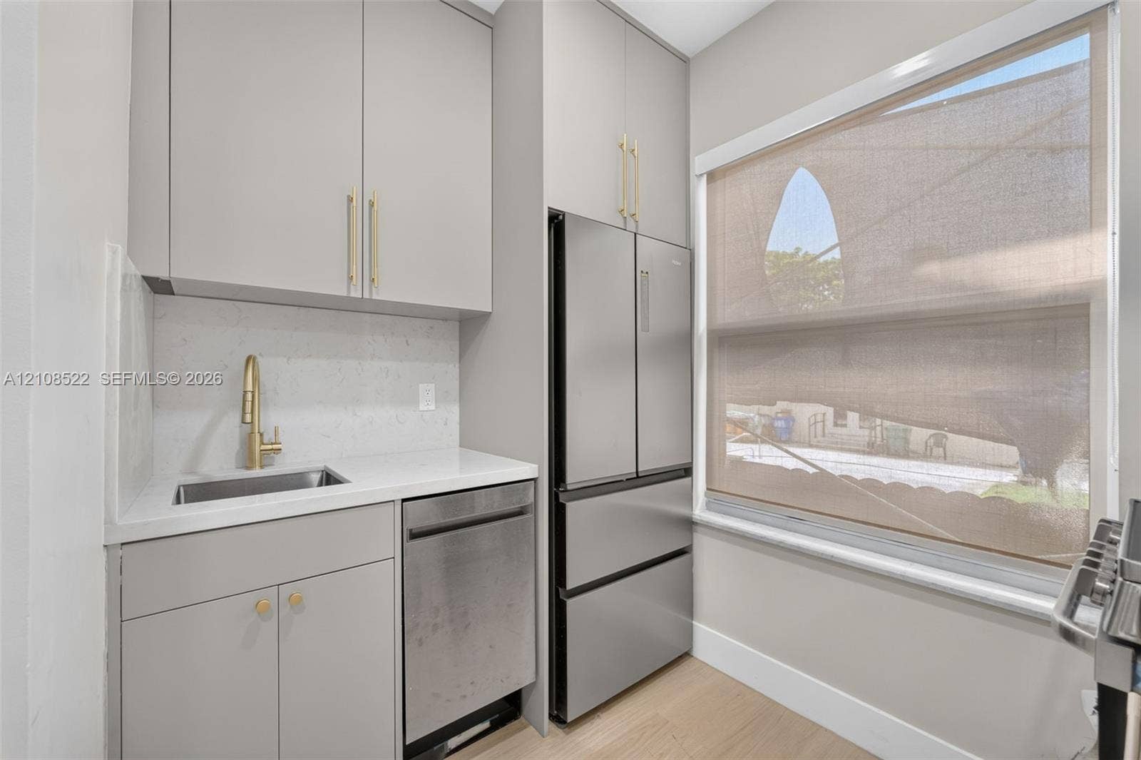 Light grey cabinetry with brass-finish hardware, white stone-composite countertop, stainless steel dishwasher, integrated refrigerator, and undermount sink with high-arc gold-finish faucet