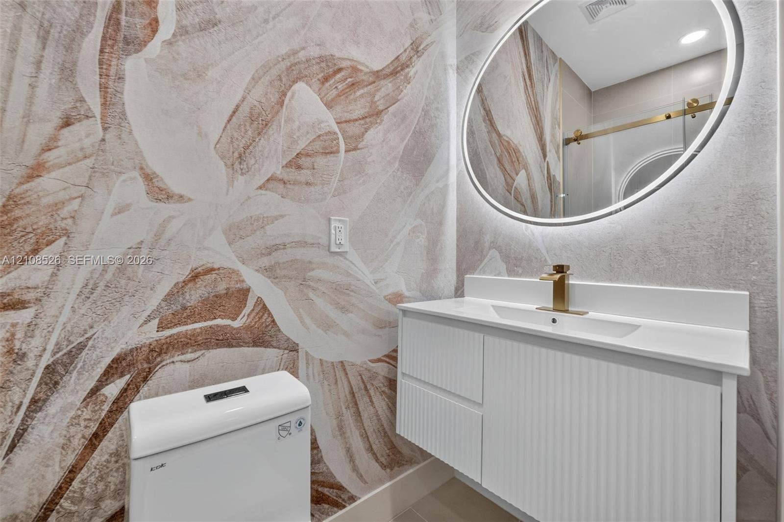 Floating white vanity with textured cabinetry, integrated countertop, brushed gold faucet, and circular LED-backlit mirror against a patterned wall treatment