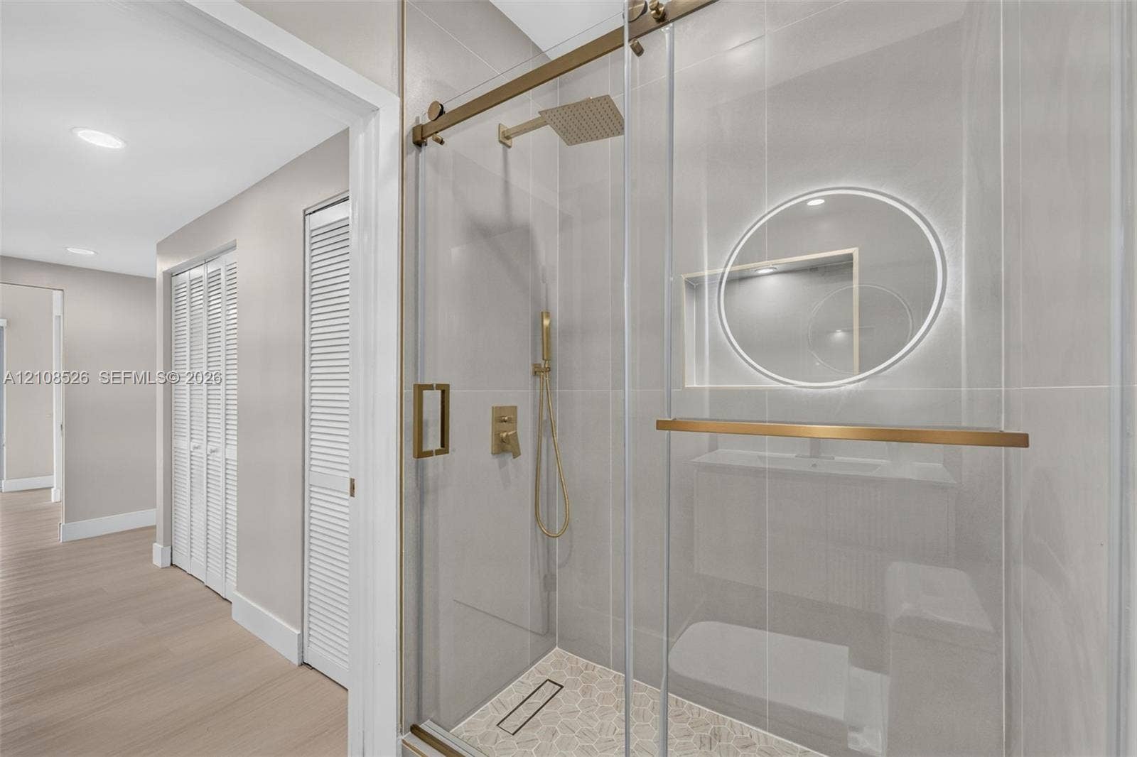 Walk-in shower with floor-to-ceiling tile, brass-finish fixtures, rainfall showerhead, and sliding glass door, paired with a circular backlit mirror