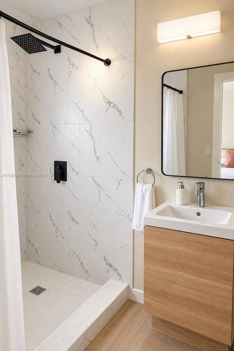 1124 N 18th Avenue N, Lake Worth, FL 33460 Walk-in shower featuring large-format tile with veining, matte black rainfall shower head, wood-finish vanity with integrated sink, rectangular wall mirror with black frame, and wall-mounted light fixture