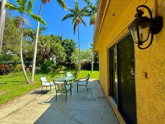 1124 NE 7th Street, Hallandale Beach, Hallandale Beach, FL 33009 Exterior patio featuring a concrete surface, stucco siding, sliding glass doors, and a wall-mounted coach light, overlooking a lawn with mature palm trees