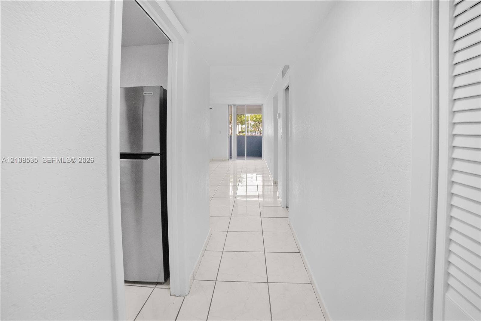 Hallway featuring neutral tile flooring, white walls, louvered closet doors, and a stainless steel refrigerator