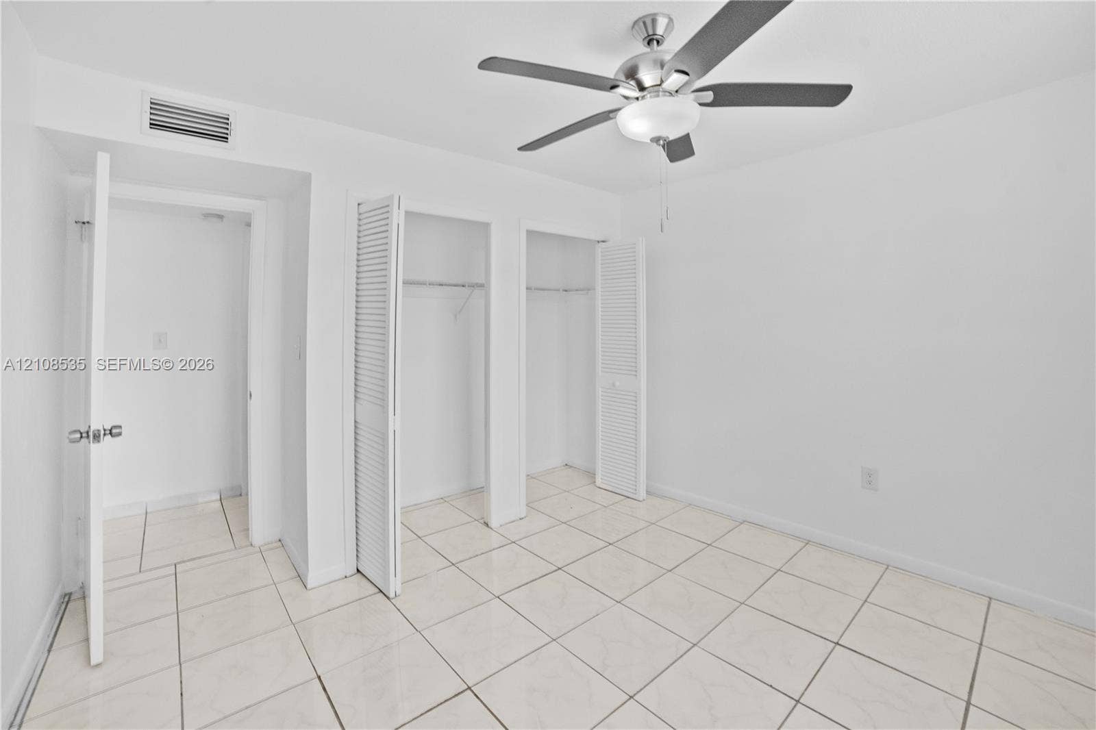 Room featuring light-toned tile flooring, neutral wall finishes, dual closets with louvered doors, and a ceiling-mounted light fixture with fan