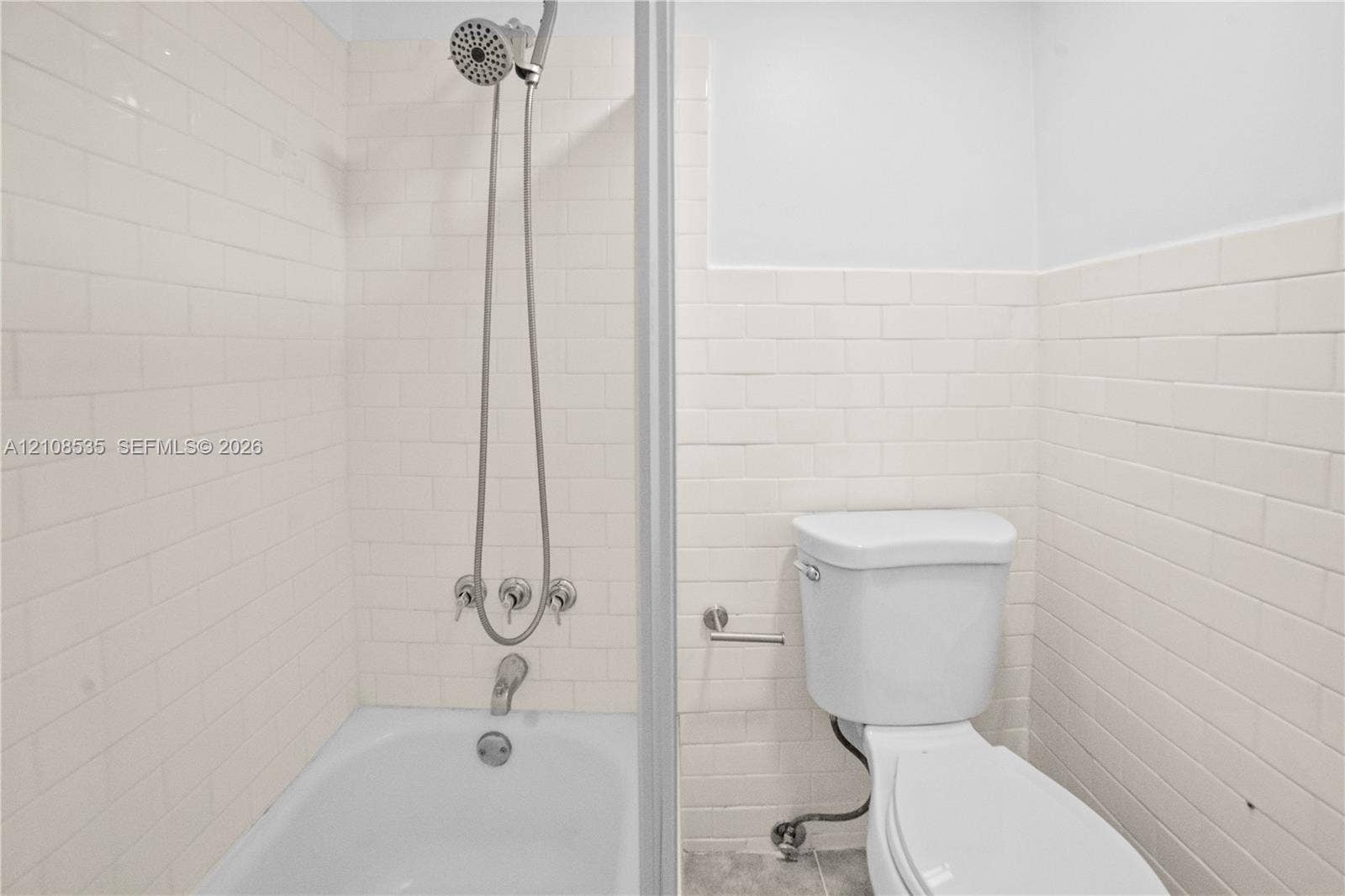 Bathroom featuring white subway tile wall surround, chrome wall-mounted shower fixture with handheld sprayer, built-in bathtub, and porcelain toilet