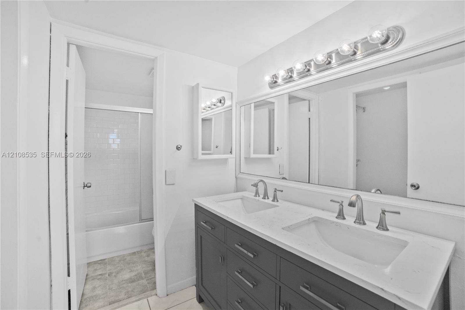 Dual-sink vanity with a solid-surface countertop, cabinetry featuring matte-finish hardware, large wall-mounted mirror, multi-bulb vanity lighting, and tub-shower combination with glass enclosure