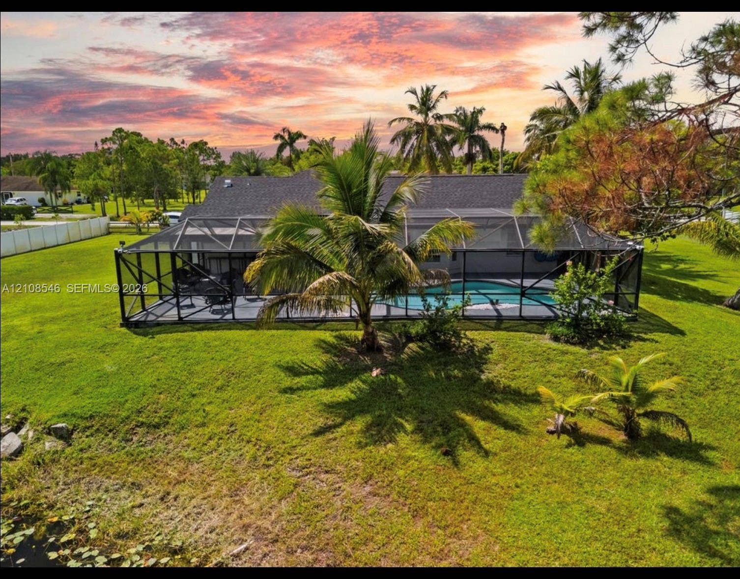 Screened-in pool enclosure, manicured lawn, mature palm trees, shingle roof, and expansive rear lot