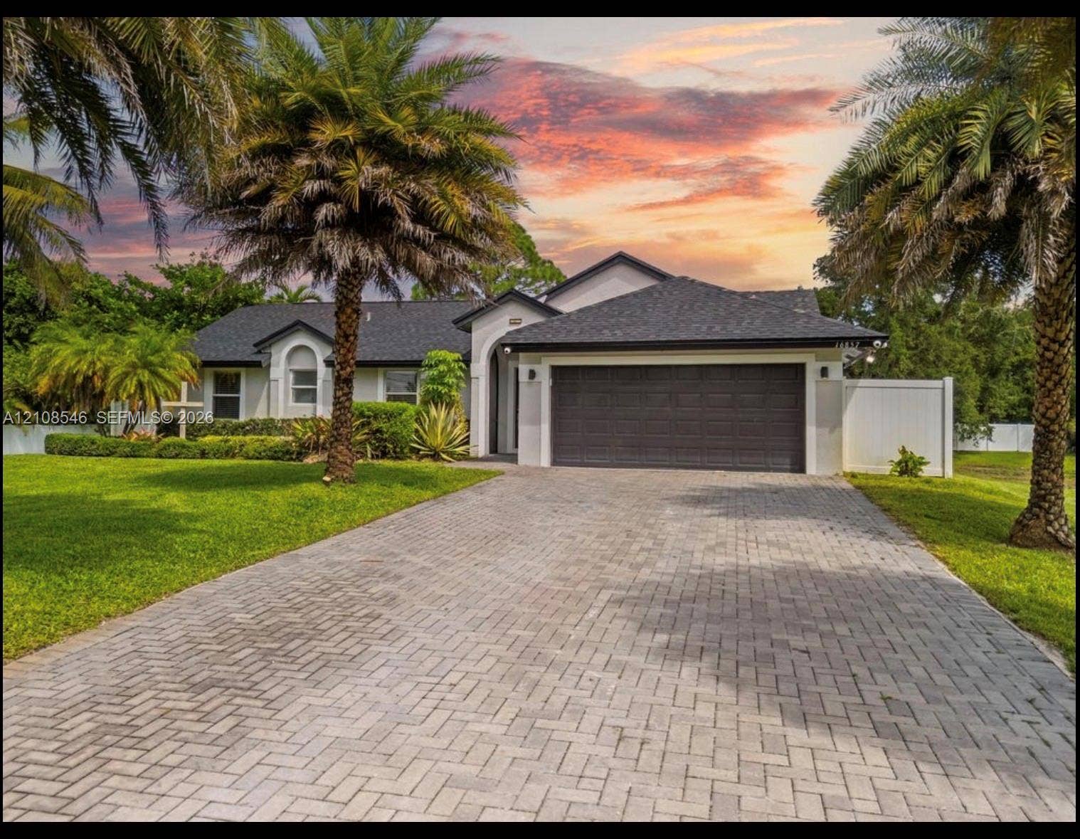 Single-story exterior featuring a shingled roof, paver driveway, attached two-car garage, manicured lawn, and mature palm trees