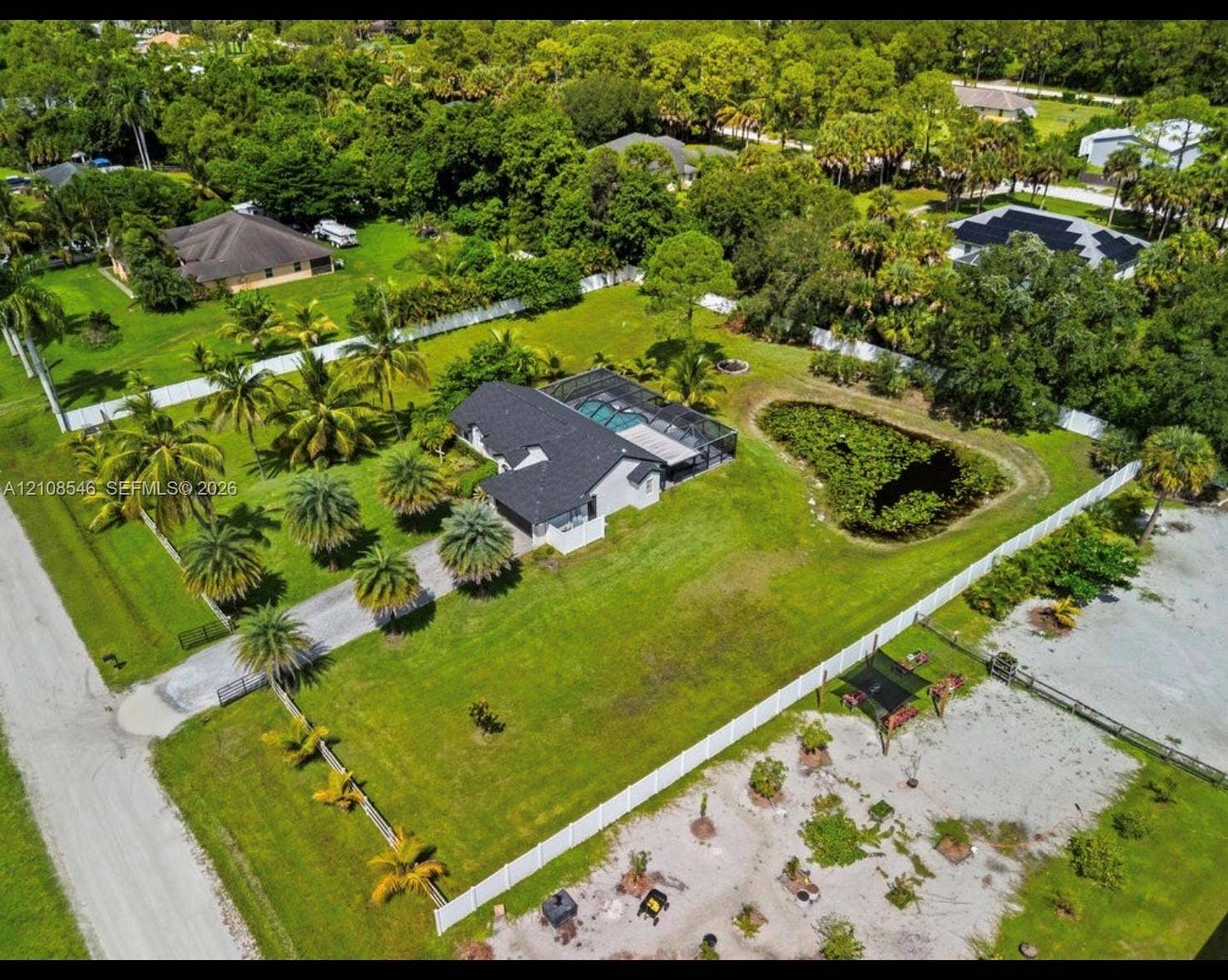 Aerial perspective of a sprawling lot featuring a residence with a screen-enclosed pool, a private pond, multiple palm trees, and full-perimeter vinyl fencing