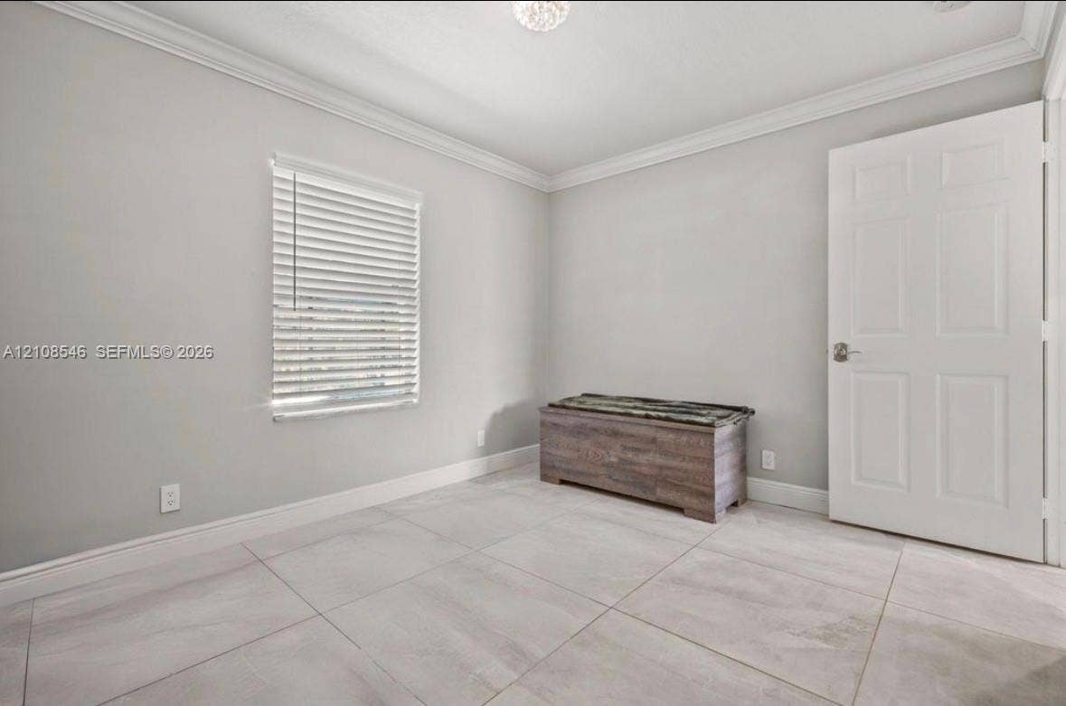 Neutral-toned interior space featuring large-format ceramic tile flooring, crown molding, a multi-panel interior door, and horizontal window blinds