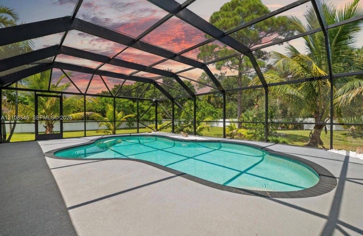 Free-form swimming pool with a tile surround, encased in a screened-in aluminum lanai structure featuring a concrete deck and lush tropical landscaping