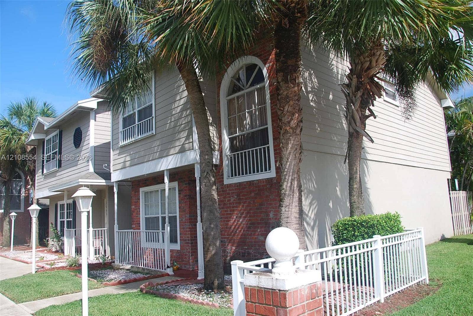 Multi-story exterior featuring a red brick and horizontal siding facade, arched second-story windows, metal balcony railings, and mature palm trees