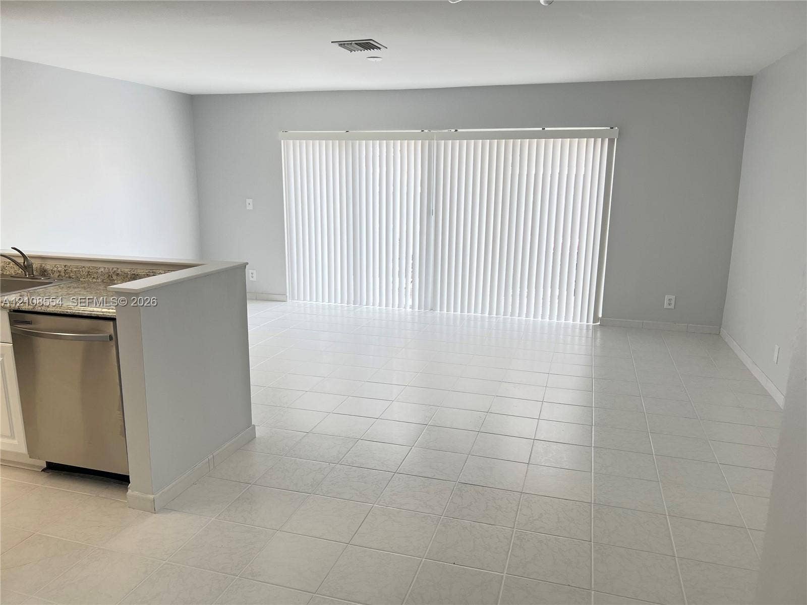 Neutral-toned interior featuring ceramic tile flooring, kitchen peninsula with stone-finish countertop, stainless steel dishwasher, and sliding glass doors with vertical blinds
