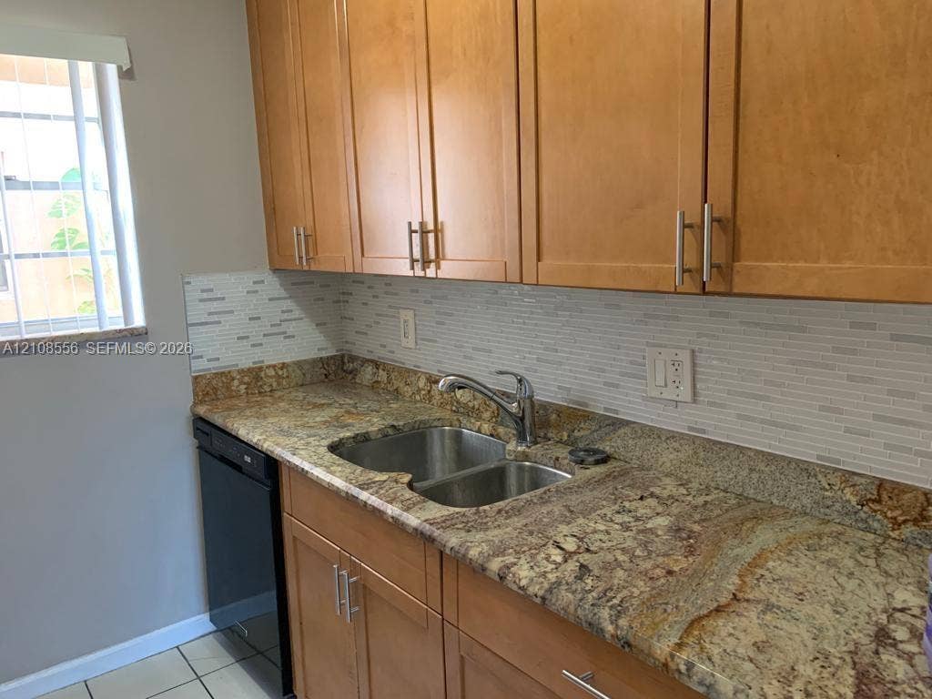600 SW 2nd Avenue, #137, Boca Raton, FL 33432 Kitchen featuring warm-toned wood cabinetry with bar-pull hardware, stone-surface countertops, a double-basin stainless steel sink, a mosaic-tile backsplash, and a built-in black dishwasher