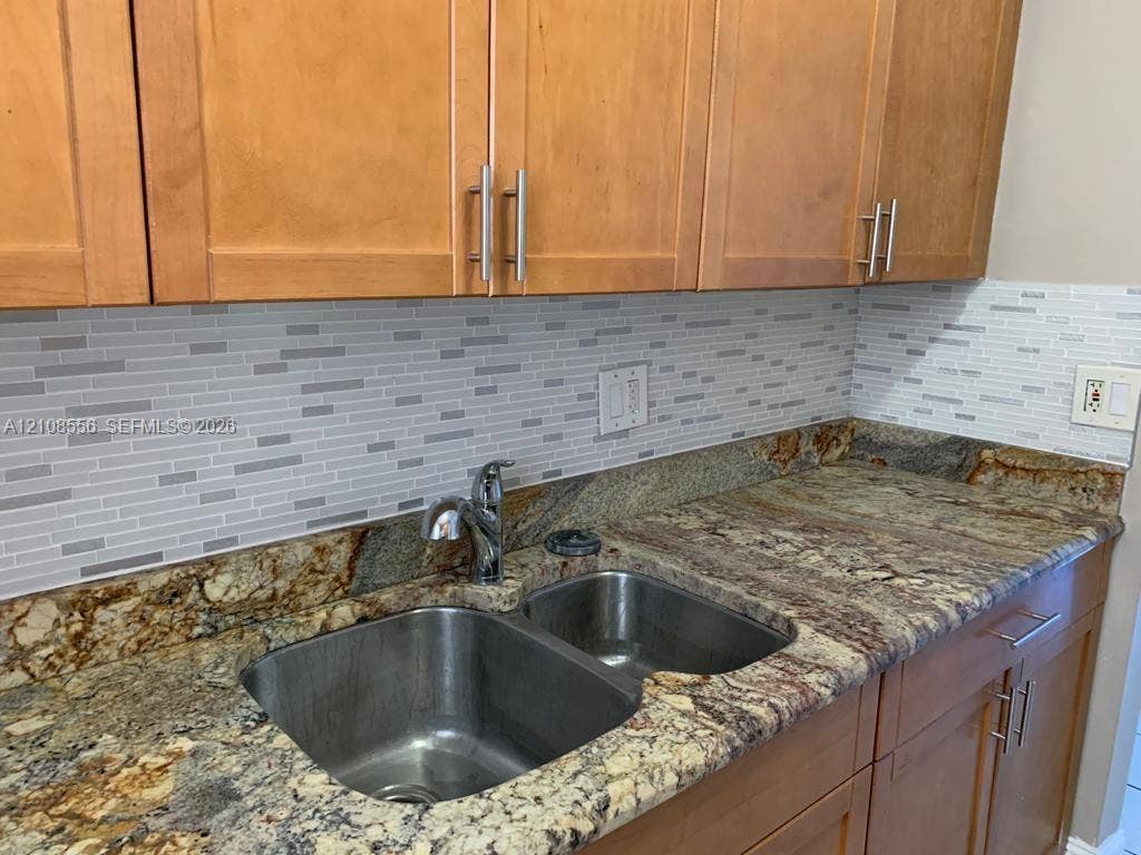 600 SW 2nd Avenue, #137, Boca Raton, FL 33432 Natural stone countertop with an integrated double-basin stainless steel sink, polished chrome faucet, linear mosaic tile backsplash, and light-tone wood-finish cabinetry featuring brushed metal hardware