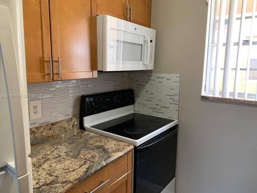 600 SW 2nd Avenue, #137, Boca Raton, FL 33432 Kitchen featuring wood-finish cabinetry with bar pulls, stone-patterned countertops, a tiled linear mosaic backsplash, and a built-in over-the-range microwave